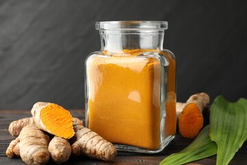 Aromatic turmeric powder in glass jar, fresh roots and leaves on wooden table, closeup