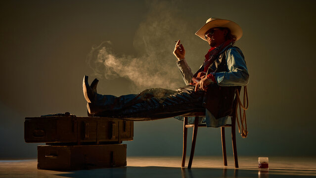 Handsome, brutal man, western cowboy sitting on chair, wearing hat, bandana, and demin jacket, reclining and smoking cigarette on dark background with warm lightning. Whiskey glass standing on floor