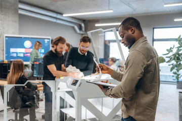 African American man is standing in front of his colleagues. People are in the office by the 3d printer