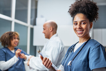Smiling Nurse in Scrubs Holding a Clipboard in a Busy Healthcare Setting with Colleagues...
