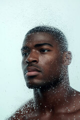Close-up of a Black man looking through a wet glass window, deep in thought
