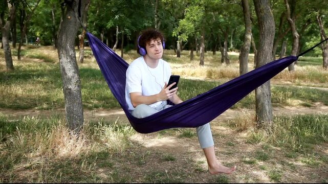 A young man enjoys relaxing in nature in a hammock, listening to music through headphones. A great moment to advertise relaxation, music and convenient accessories.