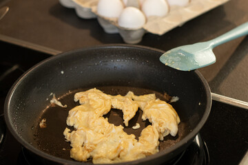 Close up of scrambled omelet egg in a black pan with a blue spatula, blurred background with white eggs 