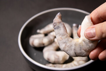 Close up of a hand holding a big frozen raw tiger prawn, blurred background with more shrimps in a grey bowl 