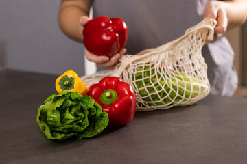 Close up of female hands putting fresh seasonal vegetables out of an eco net shopper bag on grey kitchen table
