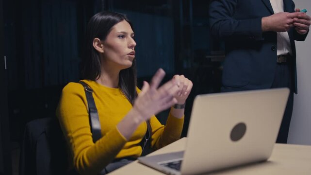 Confident young businesswoman in a yellow sweater actively discussing ideas during a late-night office meeting, using expressive gestures. Concept of workplace communication and teamwork