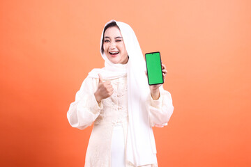 Fototapeta premium A cheerful Muslim woman in hijab holds up a smartphone with a green screen, giving a thumbs up, and smiling. isolated on a orange background.
