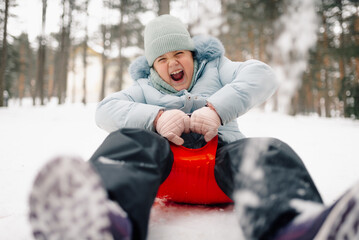 Excited girl sliding downhill on red plastic sled in snowy forest