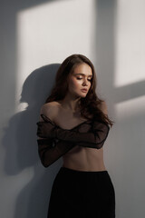 young woman stands elegantly near a wall, draped in sheer fabric that accentuates her figure. Sunlight creates gentle shadows, enhancing the artistic vibe of the indoor atmosphere