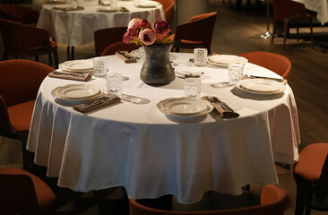 A round table with a crisp white tablecloth, set with plates, cutlery, glasses, and a floral centerpiece, ready for diners
