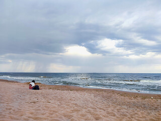 Obraz premium Woman sitting on sandy beach on seashore near sea with waves during rain. Dark storm clouds, rain and sea surface at sunset. Nature. Natural background. Stormy weather. Storm at sea.