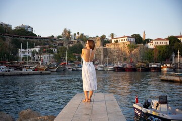 Fototapeta premium A stylish woman in a backless summer dress and sunglasses stands on a dock, gazing at the sunset over a picturesque harbor with boats and historic buildings.