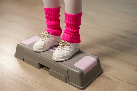 Close-up view of person's feet in white sneakers and pink leg warmers exercising on aerobic step platform indoors during workout session