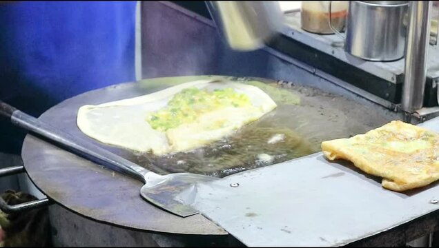 A vendor prepares an martabak telur or egg, an Indonesian specialty. Fried in hot oil on a flat pan.And use a spatula to cook .Focus on food.blurred,shake video,exposure,nighty noise
