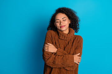 Young woman in a brown sweater embracing herself against a blue background, feeling cozy and content.