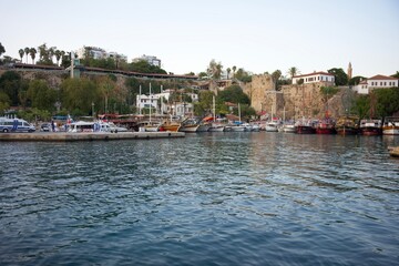 Obraz premium A scenic Mediterranean harbor with various boats docked along the waterfront, historic stone walls, lush greenery, and a coastal town backdrop. Antalya. Turkey 