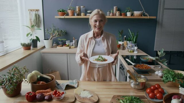 Medium portrait of happy mid-aged Caucasian woman standing ay wooden kitchen counter with white plate of beautifully served spaghetti Bolognese and smiling at camera