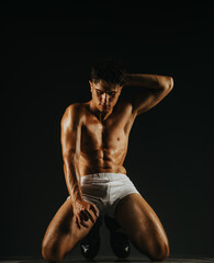 Muscular man dressed in minimal attire in a kneeling pose, highlighting physical fitness,...