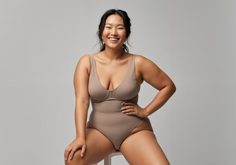 Body positive asian model smiling and posing in beige body suit, sitting on white stool in front of gray background