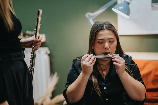 A woman passionately plays a harmonica while a second person stands holding a flute. The scene occurs indoors with a background featuring a green wall, soft lighting, and cozy furnishings. - Powered by Adobe