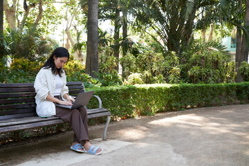 Fototapeta premium Japanese businesswoman using laptop while teleworking on a park bench, enjoying the fresh air and nature