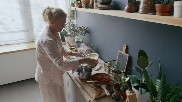 Medium shot of mid-aged Caucasian woman rinsing pasta in colander after cooking, preparing homemade spaghetti Bolognese in cozy kitchen setting
