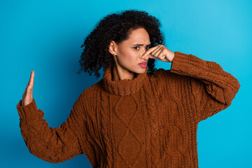 Young woman pinching her nose in disgust and raising her hand, wearing a brown sweater with blue...