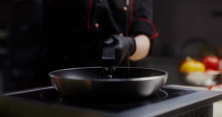 A woman in a chef's uniform with disposable black gloves on her hands pours oil on a frying pan standing on the stove, a close-up of her hands