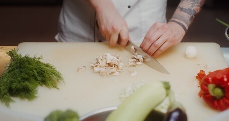 A man in a chef's uniform cuts mushrooms and then puts them in a frying pan standing on the stove, close-up of his hands
