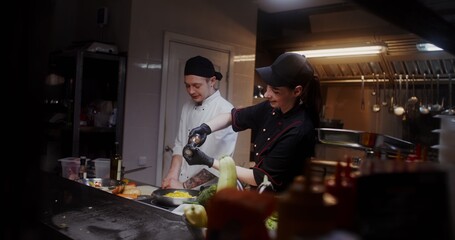 A man and a woman in a chef's uniform talk and smile while cooking in the restaurant kitchen together