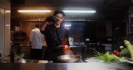 A woman in a chef's uniform adds food to a frying pan with fire while cooking in a restaurant kitchen