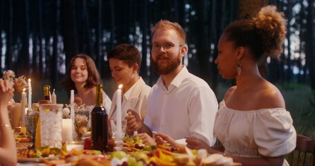 Guests laugh and talk while sitting at a beautifully served festive table in the open air. A man lights candles