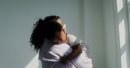 An African-American woman dressed in pastel-colored clothes holds her little daughter in her arms and strokes her back