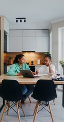 Mom helps her daughter do her homework, the girl writes in a notebook sitting at the kitchen table in front of an open laptop