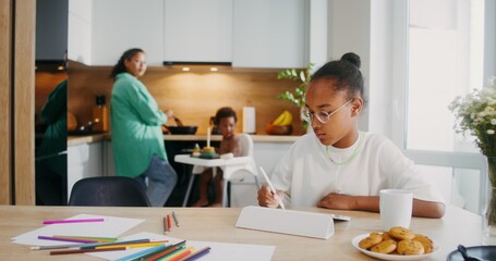 A girl draws with a stylus on a tablet while sitting at the kitchen table at home. Her family in...