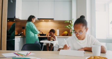 A girl draws with a stylus on a tablet while sitting at the kitchen table at home. Her family in...