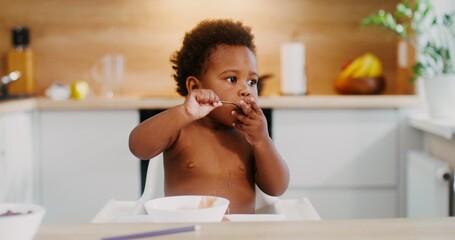 A baby with an African-American appearance with an afropric eats fruit puree with a spoon, while...