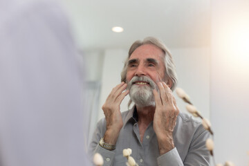 Mature Caucasian senior man looking in mirror touching the moustache. Cheerful handsome senior man looking at himself while standing in front of the mirror