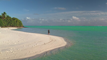 Enjoying a leisurely stroll along a pristine beach, a young woman soaks up the sun and the...