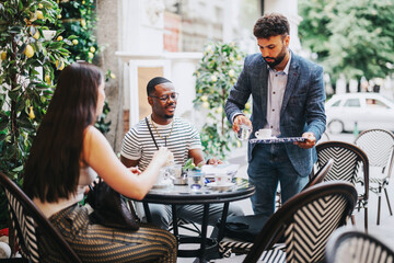 A group of friends relax at a stylish outdoor cafe while a waiter serves coffee. The scene captures a moment of leisure and socializing in a vibrant urban setting.