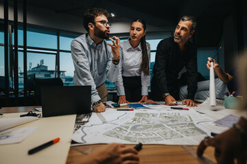 Business professionals engage in a strategic discussion, analyzing blueprints and data plans around...