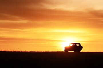 Silhouetted against a vibrant sunset, an off-road vehicle navigates a vast grass field, evoking feelings of freedom and adventure in a stunning landscape