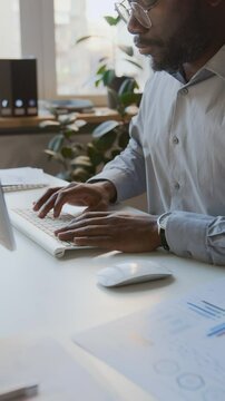Medium vertical shot of African American male manager or business analyst typing on computer keyboard in office, while working on quarterly presentation with financial graphs