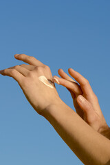 Vertical image of female hands applying moisturizer to skin against blue sky background. Concept of texture and swatch of body care product, spa and aesthetics