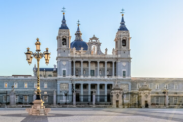 Almudena Cathedral in Madrid, Spain during sunset, with a beautiful ornate street lamp in the foreground. The soft golden light highlights the neoclassical facade of this iconic religious landmark