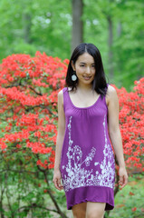 Fototapeta premium Beautiful young Asian woman in purple sundress walks away from bright and blooming shrub in background