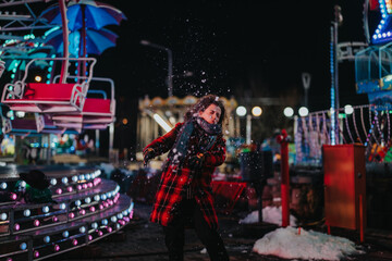 Obraz premium A woman enjoying herself on a snowy night at a brightly lit amusement park. The scene captures her playful and joyful mood alongside the festive colors of the winter season.