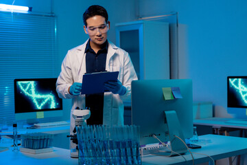 Asian male scientist wearing lab coat and protective glasses, holding clipboard and analyzing data in a modern laboratory with computers and microscopes