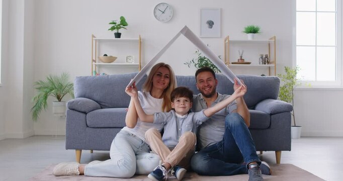 Happy family with child is sitting at home, holding a cardboard roof above heads. The scene symbolizes their dream of a new house, love, and the protection of a caring and secure home environment.