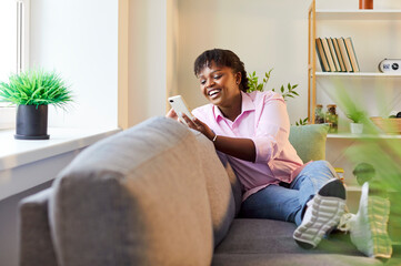 Happy african american woman at home sitting on sofa with mobile phone to enjoy chat with friend via messenger or social network online communication, application for shopping, send, read good message